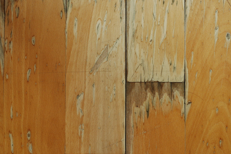 Close-up image of a section of wooden flooring in a residential or commercial space, showing multiple areas of wear, scratches, and surface damage. The flooring boards are characterized by a light to medium oak finish, with visible grain patterns and knot marks. Some sections display chipped, peeling, or stained finishes, indicating the need for professional cleaning or restoration. The surface appears uneven in places, with cracks and scuffs evident, likely caused by foot traffic or impact. The environment suggests an indoor room, possibly a living room or hallway, with natural or artificial light highlighting the condition of the wood. The image aligns with the work of professional cleaning services such as Carpet Shampoo in London, especially for clients seeking restoration of damaged wooden or laminate flooring around postcode areas in London.