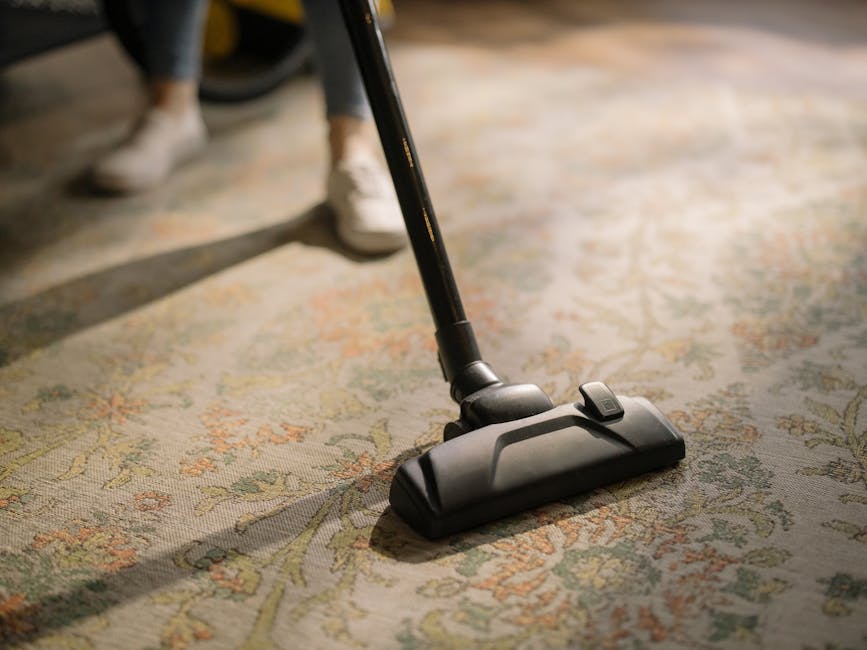 A close-up view of a person vacuuming an ornate, patterned carpet in a domestic setting, demonstrating surface cleaning with a modern black vacuum cleaner. The scene captures the vacuum head pressed f