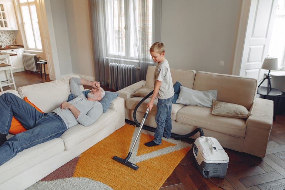 A young boy using a vacuum cleaner to clean a synthetic rug in a living room with natural light coming through large windows. An older man is lying on a light-colored fabric sofa, appearing relaxed an