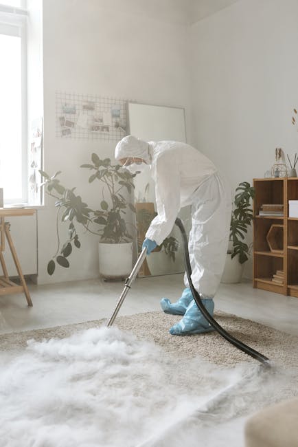 A middle-aged man with gray hair and a beard is performing deep surface cleaning in a bright living room, using a vacuum cleaner to extract dirt from a patterned yellow, white, and pink rug laid on a 